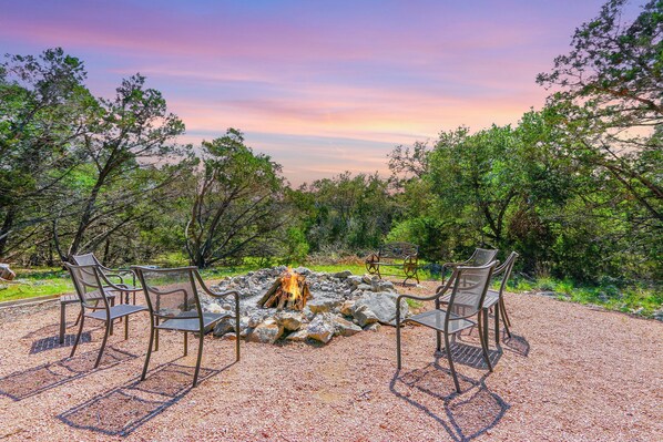 An outdoor seating area with a fire pit surrounded by chairs, set against a backdrop of trees and a colorful sky. - Rim Rock I Blanco, Log Cabin , Pool, Hot Tub, 10 acres of Nature to Explore (Blanco)