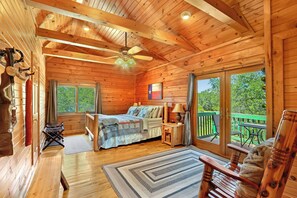 A wooden cabin with a bed, a small table, a chair, and a balcony with a view of trees.