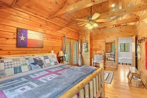 A wooden cabin interior with a bed, a ceiling fan, and a view of the bathroom. - Rim Rock I Blanco, Log Cabin , Pool, Hot Tub, 10 acres of Nature to Explore (Blanco)