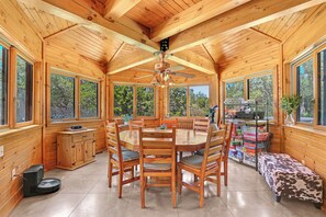 A wooden cabin interior with a round dining table, wooden chairs, a ceiling fan, and large windows offering views of the outdoors. - Rim Rock I Blanco, Log Cabin , Pool, Hot Tub, 10 acres of Nature to Explore (Blanco)