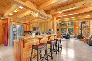A wooden cabin interior with a kitchen, bar stools, and a stone fireplace. - Rim Rock I Blanco, Log Cabin , Pool, Hot Tub, 10 acres of Nature to Explore (Blanco)