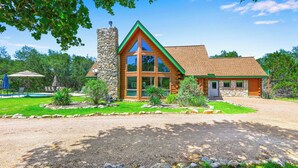 A log cabin with a stone chimney, large windows, and a well-maintained lawn. - Rim Rock I Blanco, Log Cabin , Pool, Hot Tub, 10 acres of Nature to Explore (Blanco)