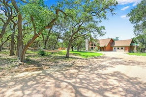 A wooden cabin with a stone foundation, surrounded by a forested area with a gravel driveway. - Rim Rock I Blanco, Log Cabin , Pool, Hot Tub, 10 acres of Nature to Explore (Blanco)