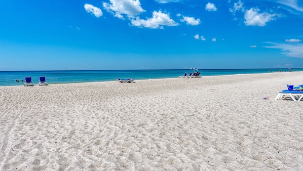 Una playa con arena blanca y agua azul cristalina. Se observan sillas de playa y sombrillas azules.