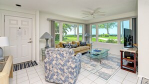 Un salón espacioso con sofá, sillón y mesa de centro, ventanas amplias con vistas a zonas verdes, un ventilador de techo y televisor.