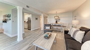 A spacious living room with a dark brown sofa, a wooden coffee table, and a dining area with a chandelier. - Unit #117 Sand Cay Beach Resort Gulf Front (Longboat Key)