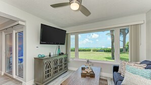 A modern living room with a flat-screen TV, a wooden coffee table, a gray sofa with patterned cushions, and a view of a green landscape through large windows. - Unit #117 Sand Cay Beach Resort Gulf Front (Longboat Key)
