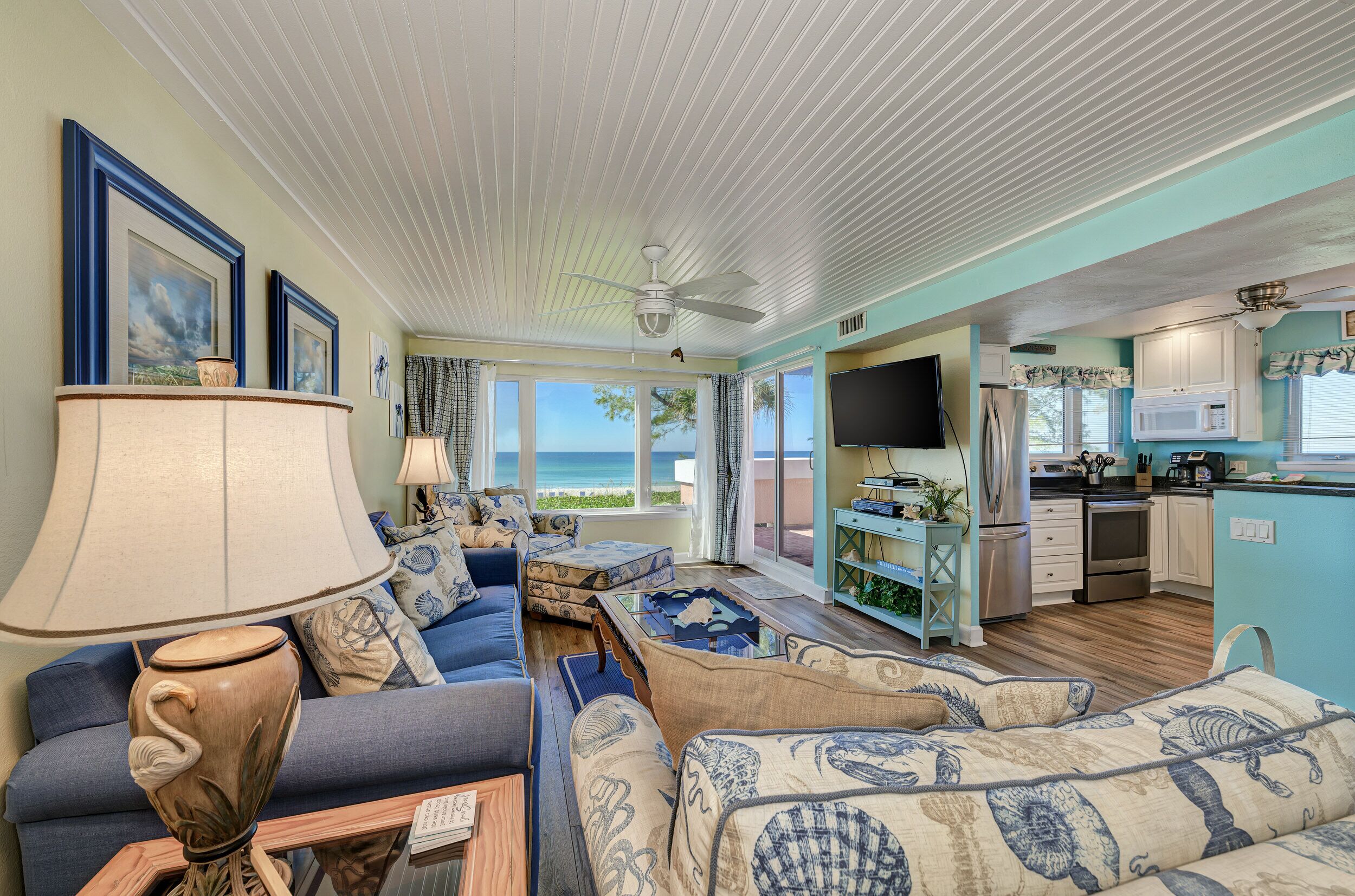 A spacious living room with a large sofa, a coffee table, and a TV. There is a kitchen area with a microwave, refrigerator, and cabinets. The room has large windows with a view of the ocean.
