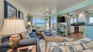 A spacious living room with a large sofa, a coffee table, and a TV. There is a kitchen area with a microwave, refrigerator, and cabinets. The room has large windows with a view of the ocean. - Unit #217 Sand Cay Beach Resort Gulf Front (Longboat Key)