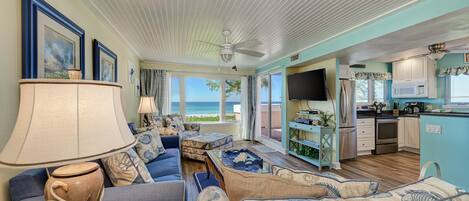A spacious living room with a large sofa, a coffee table, and a TV. There is a kitchen area with a microwave, refrigerator, and cabinets. The room has large windows with a view of the ocean.