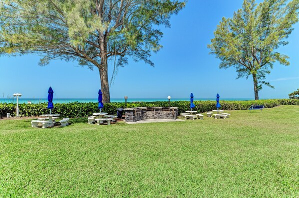 A grassy outdoor area with picnic tables and blue umbrellas, a stone barbecue area, and a view of the sea.