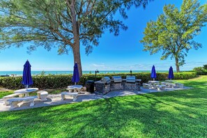 An outdoor seating area with stone tables and benches, surrounded by trees and a view of the ocean.