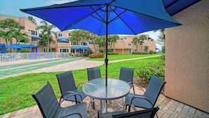 A patio area with a round table and chairs, a large blue umbrella, and a view of a residential complex with multiple buildings and a green lawn.