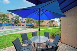 A patio area with a round table and chairs, a large blue umbrella, and a view of a residential complex with multiple buildings and a green lawn. - Unit #107 Sand Cay Beach Resort Pool View (Longboat Key)