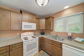 Une cuisine avec des meubles de rangement en bois, une cuisinière blanche, un four à micro-ondes, un évier et une fenêtre avec des stores.