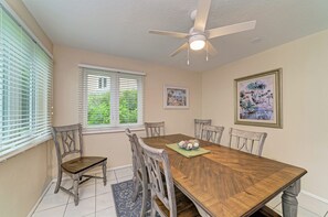 Un coin repas comprenant une table en bois, six chaises, un ventilateur de plafond et deux fenêtres équipées de stores.
