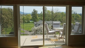 Et værelse med en skydedør, der fører ud til en terrasse med bord og stole, samt udsigt over et grønt landskab med træer og en bygning i det fjerne.