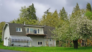 Et toetagers hus med balkon, et drivhus og et træ med hvide blomster.