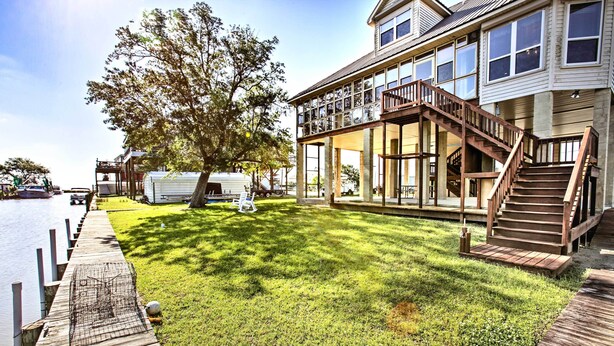A waterfront property with a wooden dock, a grassy lawn, and a two-story house with a balcony and stairs. - Private Dock w/ Lift: Waterfront New Orleans Home! (New Orleans)