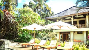A pool area with lounge chairs and umbrellas, surrounded by lush greenery and a building with a tiled roof. - Bali's Finest, Luxurious Villa Tunjung with Private Pool&Staff in Unspoiled Bali (Seririt)