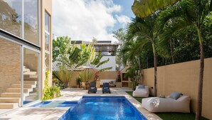Piscina moderna ao ar livre com espreguiçadeiras e um sofá branco, cercada por palmeiras e um muro de tijolos.