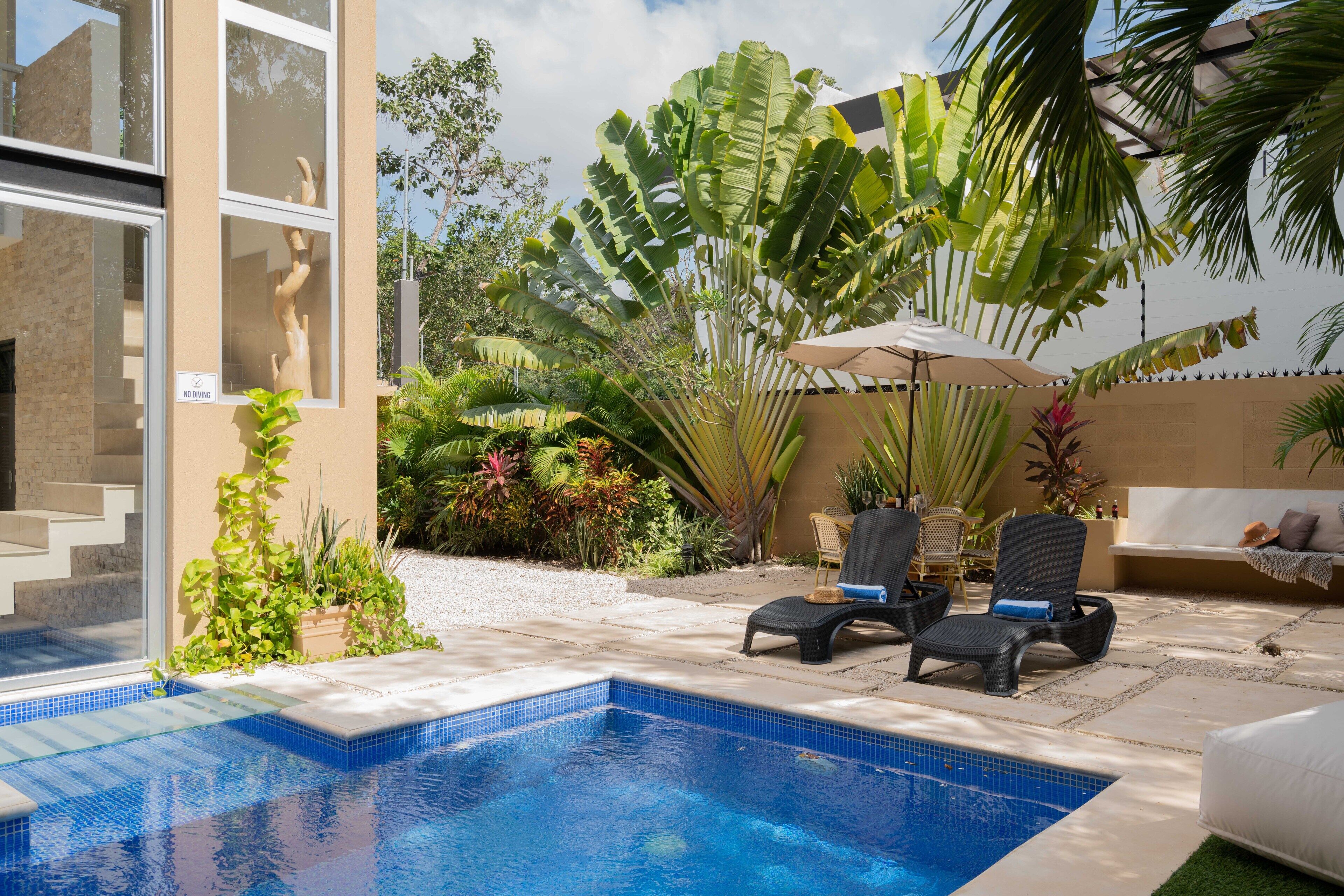 A swimming pool with lounge chairs and a covered seating area, surrounded by tropical plants and a modern building.