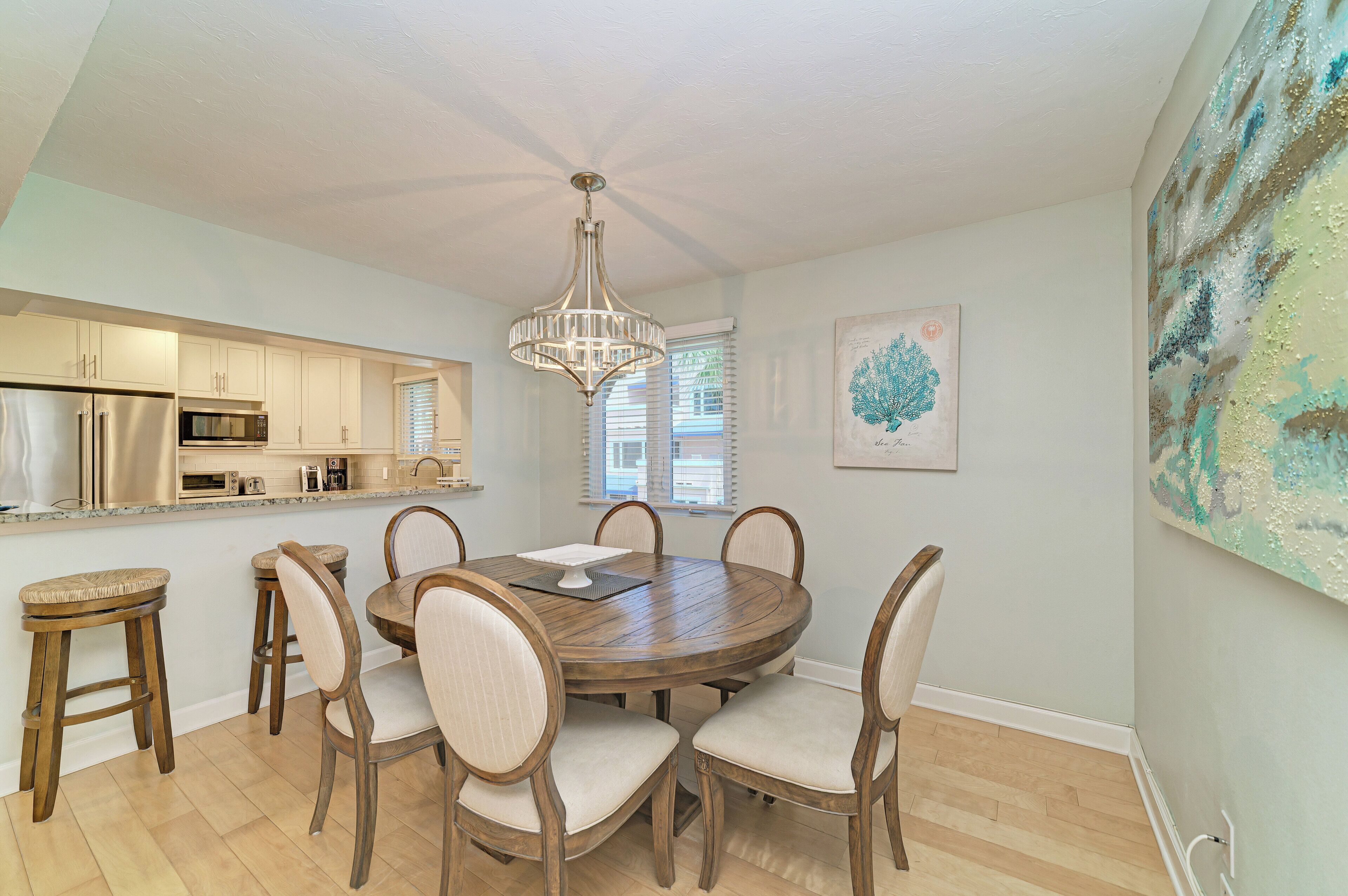 A dining area with a round wooden table, six chairs, a chandelier, a kitchen with white cabinets, and a large abstract painting on the wall.