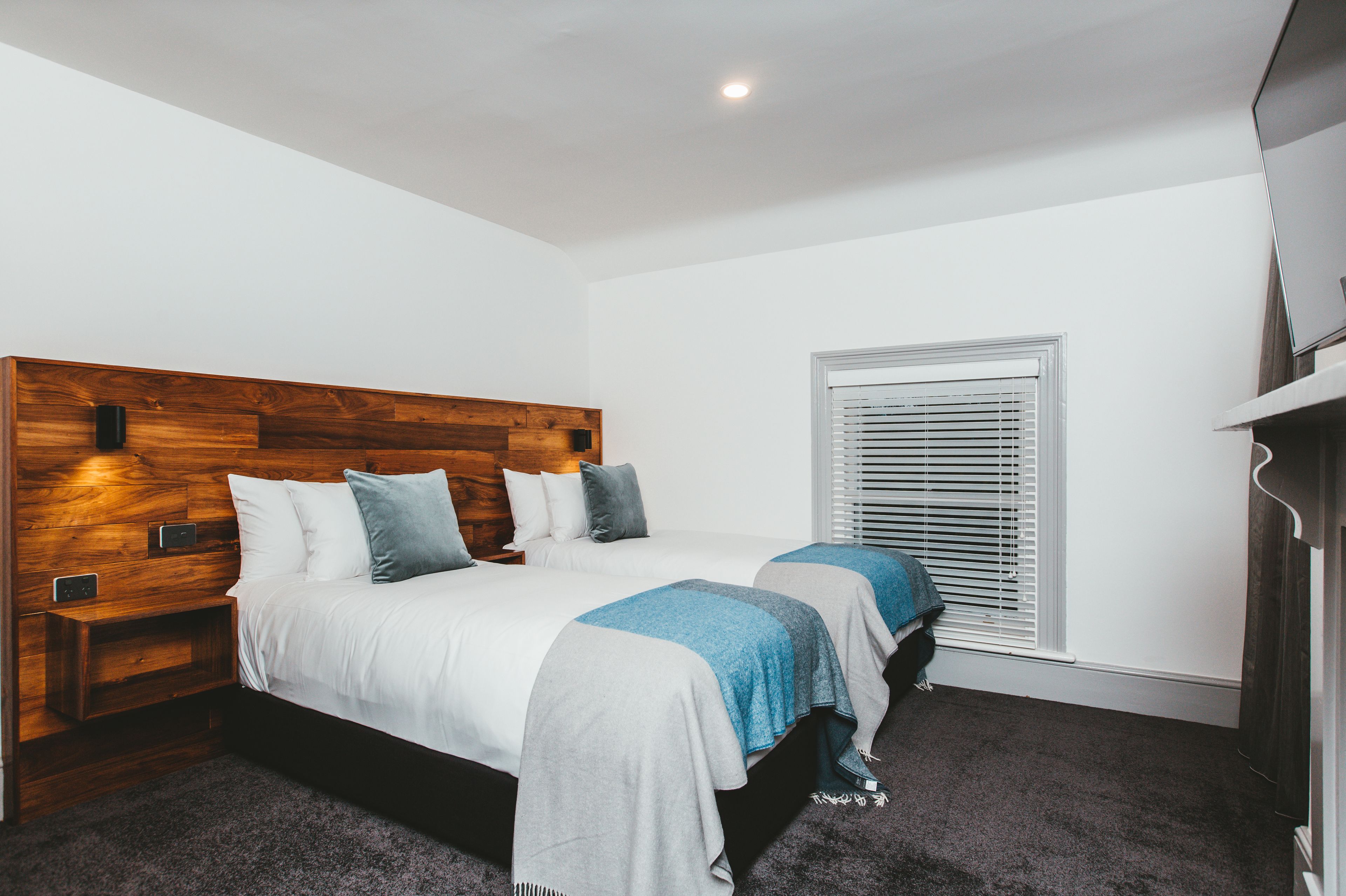 A bedroom with two beds, a wooden headboard, a window with blinds, and a dark carpet.