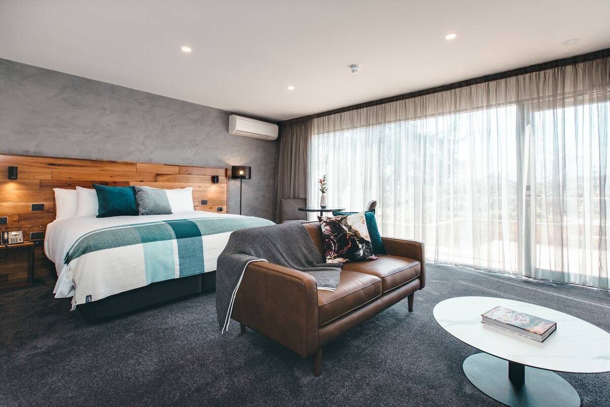 A modern hotel room with a large bed, a leather sofa, a small round table, and a large window with sheer curtains.