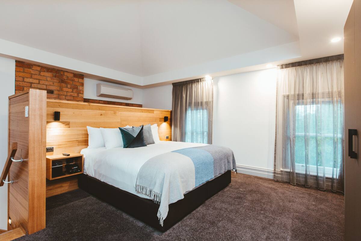 A modern hotel room with a wooden headboard, a large bed, a nightstand, a built-in wardrobe, and a large window with curtains.