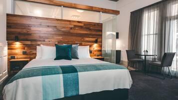 Une chambre d’hôtel moderne avec une tête de lit en bois, un grand lit recouvert d’une couverture à carreaux, deux tables de chevet avec des lampes, une petite table avec des chaises et une grande fenêtre avec des rideaux.