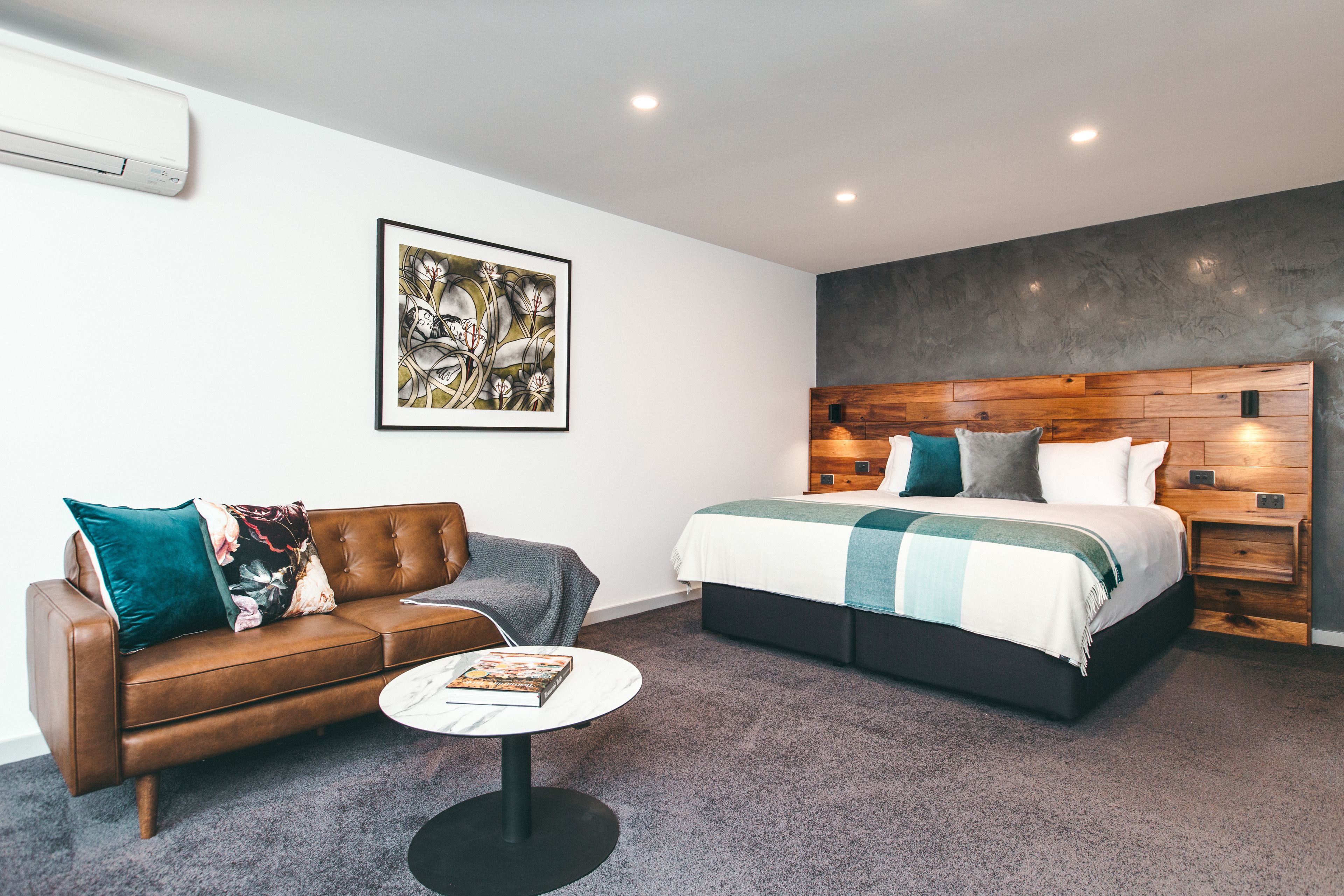 Ein modernes Hotelzimmer mit braunem Ledersofa, einem runden Couchtisch, einem großen Bett mit Holz-Kopfteil und einem gerahmten abstrakten Gemälde an der Wand.