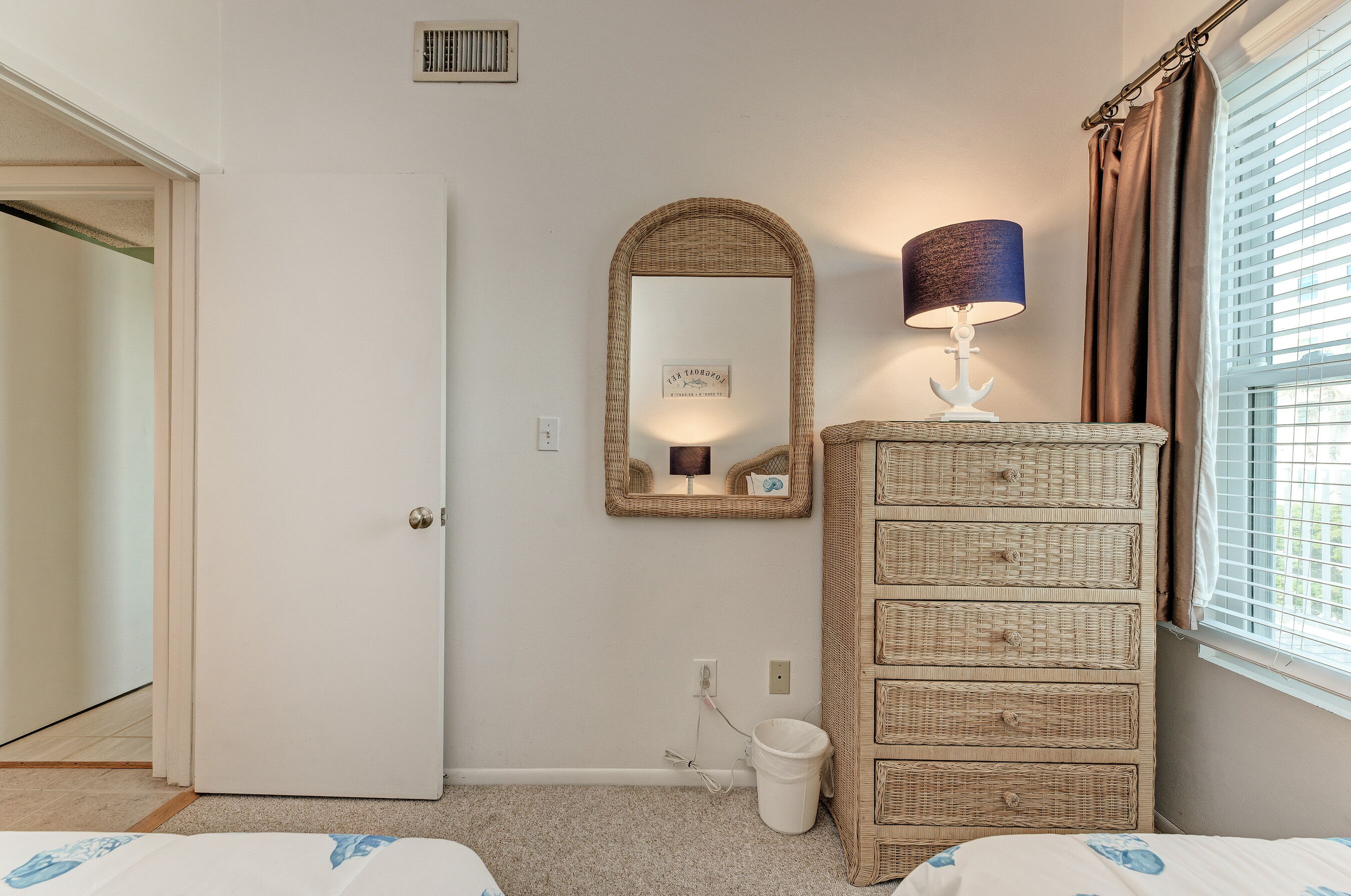 A bedroom with a white door, a wicker dresser, a mirror with a lamp, and a window with blinds.