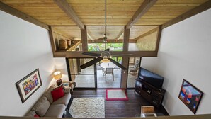 Un vaste salon doté d’un haut plafond, de poutres apparentes, d’un ventilateur de plafond, d’un téléviseur, d’un canapé et d’un coin repas visible à travers des portes vitrées.