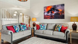 Un salon avec un canapé gris, des coussins colorés, un tableau représentant une coulée de lave et une enseigne « KONA BREWING CO ».