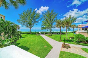 Un jardin bien entretenu, avec un sentier menant à la plage, entouré de palmiers et surplombé d’un ciel bleu azur.
