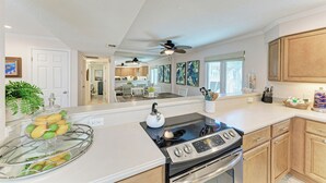 A modern kitchen with a gas stove, oven, and a glass display case with fruits. There are wooden cabinets, a ceiling fan, and artwork on the walls.
