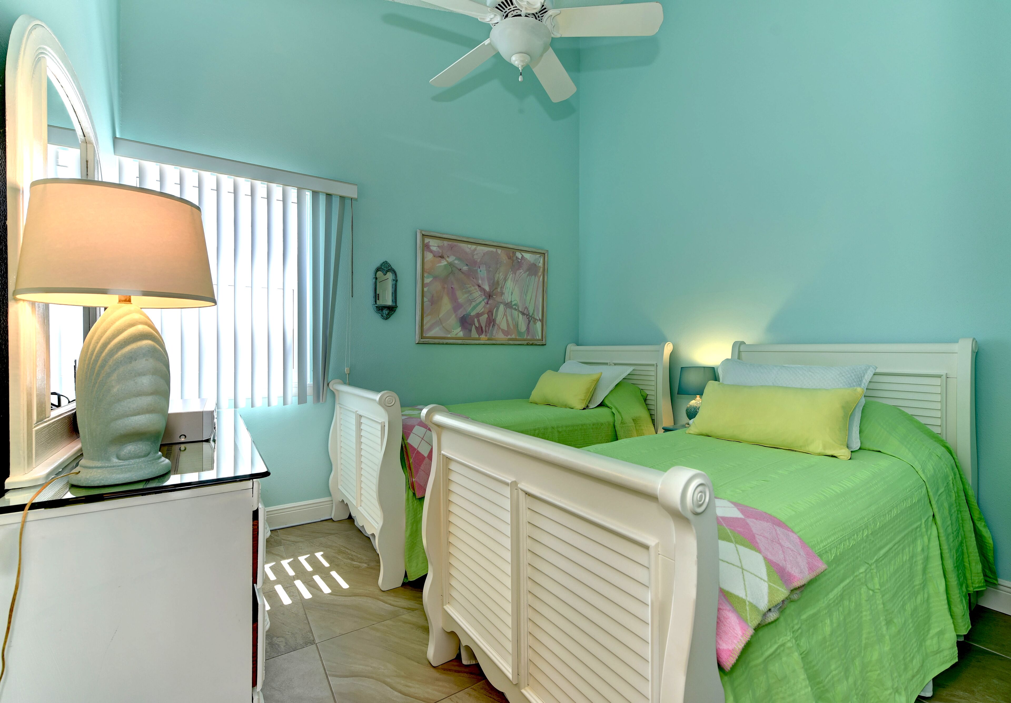 A bedroom with two beds, a ceiling fan, a bedside lamp, a radiator, and a window with blinds.