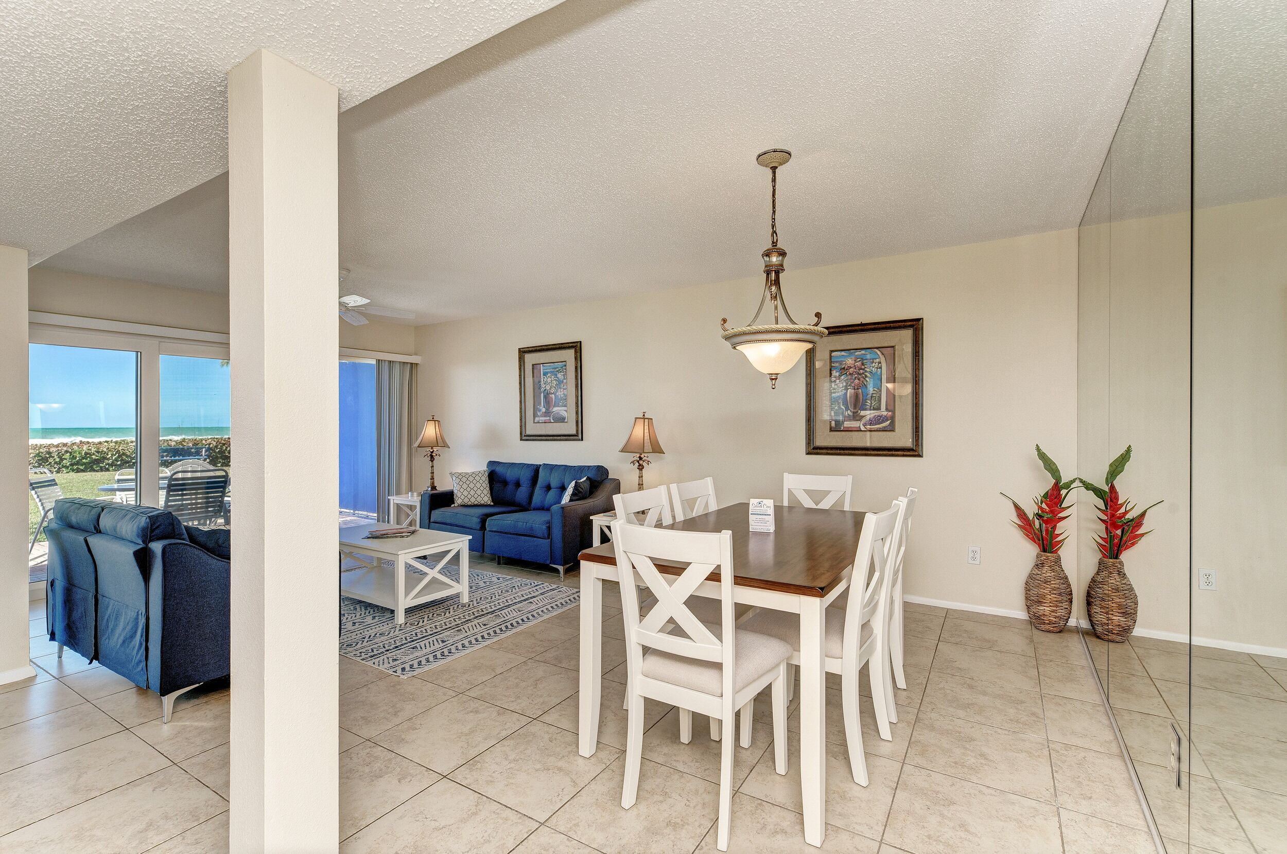 A spacious living area with a dining table, white chairs, a blue sofa, and a large window offering a view of the outdoors.