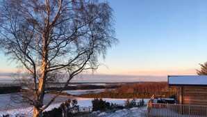 A wooden cabin with a deck, surrounded by snow-covered ground and a large tree. - 2-bed Woodland Lodge with private hot tub and expansive views. (Forres)