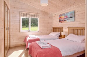 A room with two beds, a window with a view of greenery, and a painting on the wall. - 2-bed Woodland Lodge with private hot tub and expansive views. (Forres)