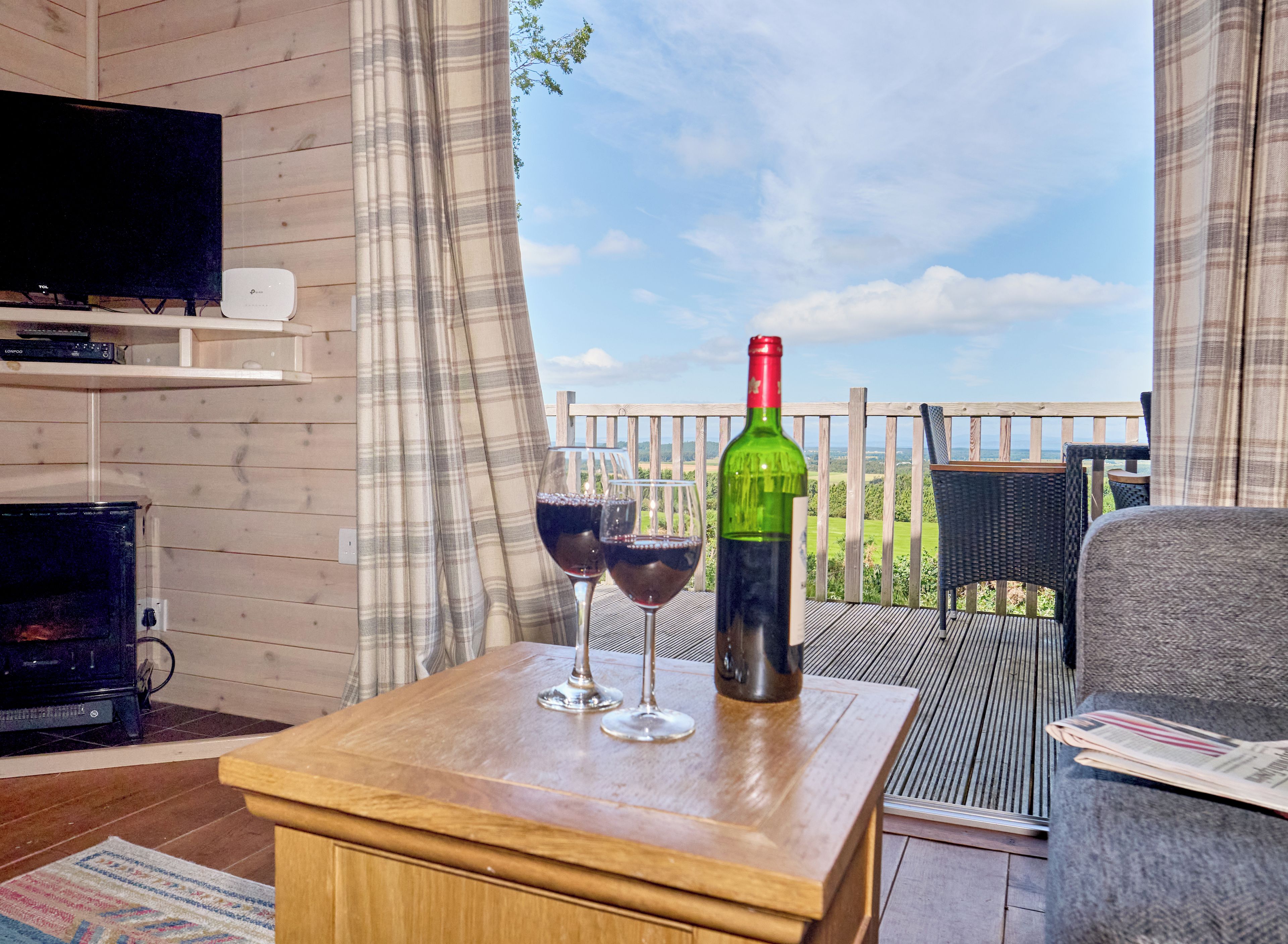 Una mesa de madera con dos copas de vino y una botella, un televisor y una silla de mimbre en un balcón con vistas al campo.