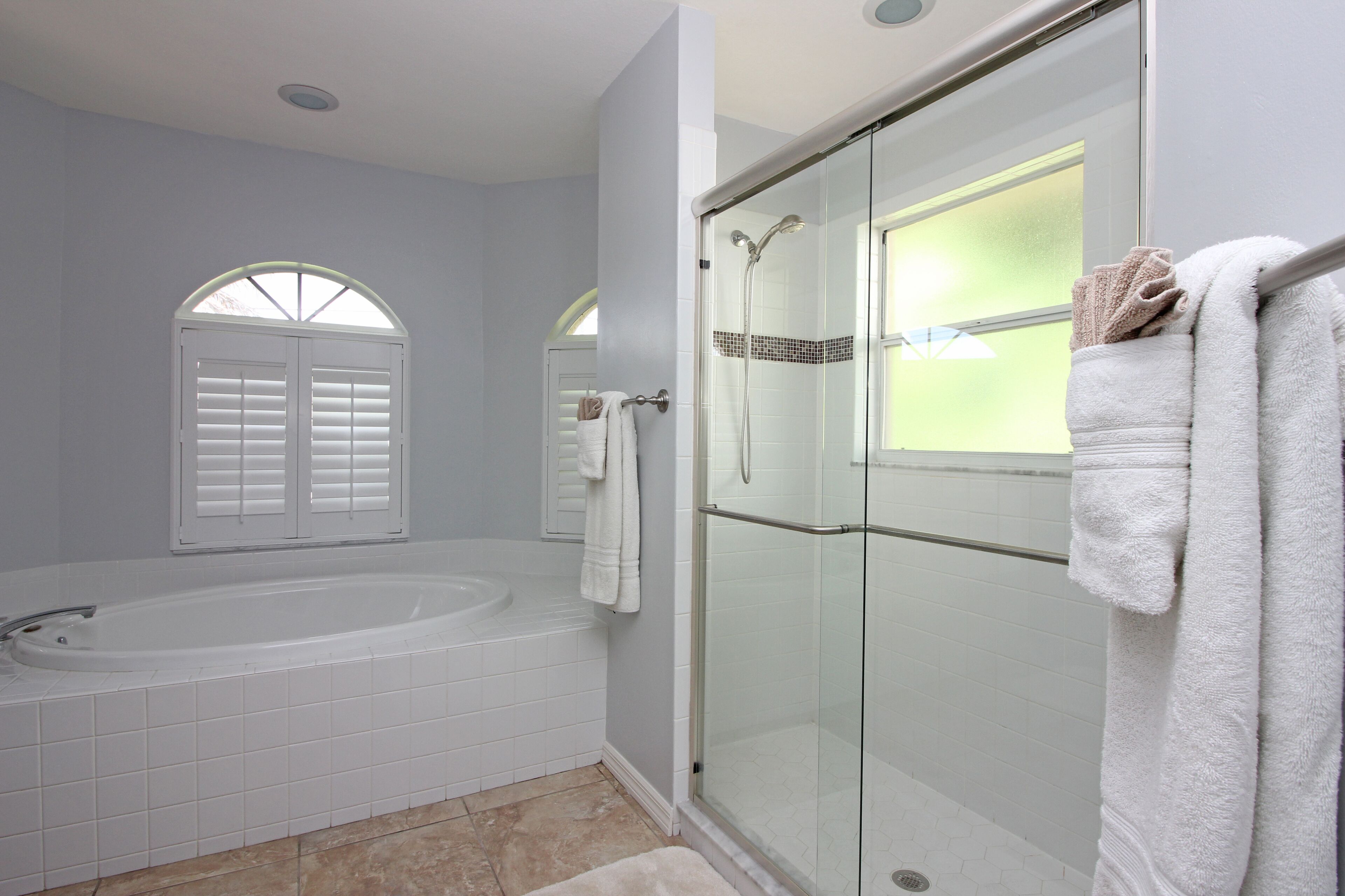 Un baño moderno con ducha envuelta en vidrio, una bañera independiente y un ventanal con contraventanas blancas.