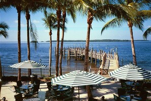Eine Terrasse am Strand mit gestreiften Sonnenschirmen, Palmen und einem Holzsteg, der ins Wasser führt.