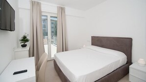 Habitación de hotel con cama, escritorio, silla, televisor y balcón con cortinas.