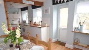 A dining area with a table set for four, a kitchen in the background, and a window with a view of a brick wall. - Old Saltys Cottage in Westward Ho with stunning sea views (Northam)