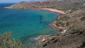 Paisagem costeira com mar azul límpido, terreno rochoso e vegetação rala.
