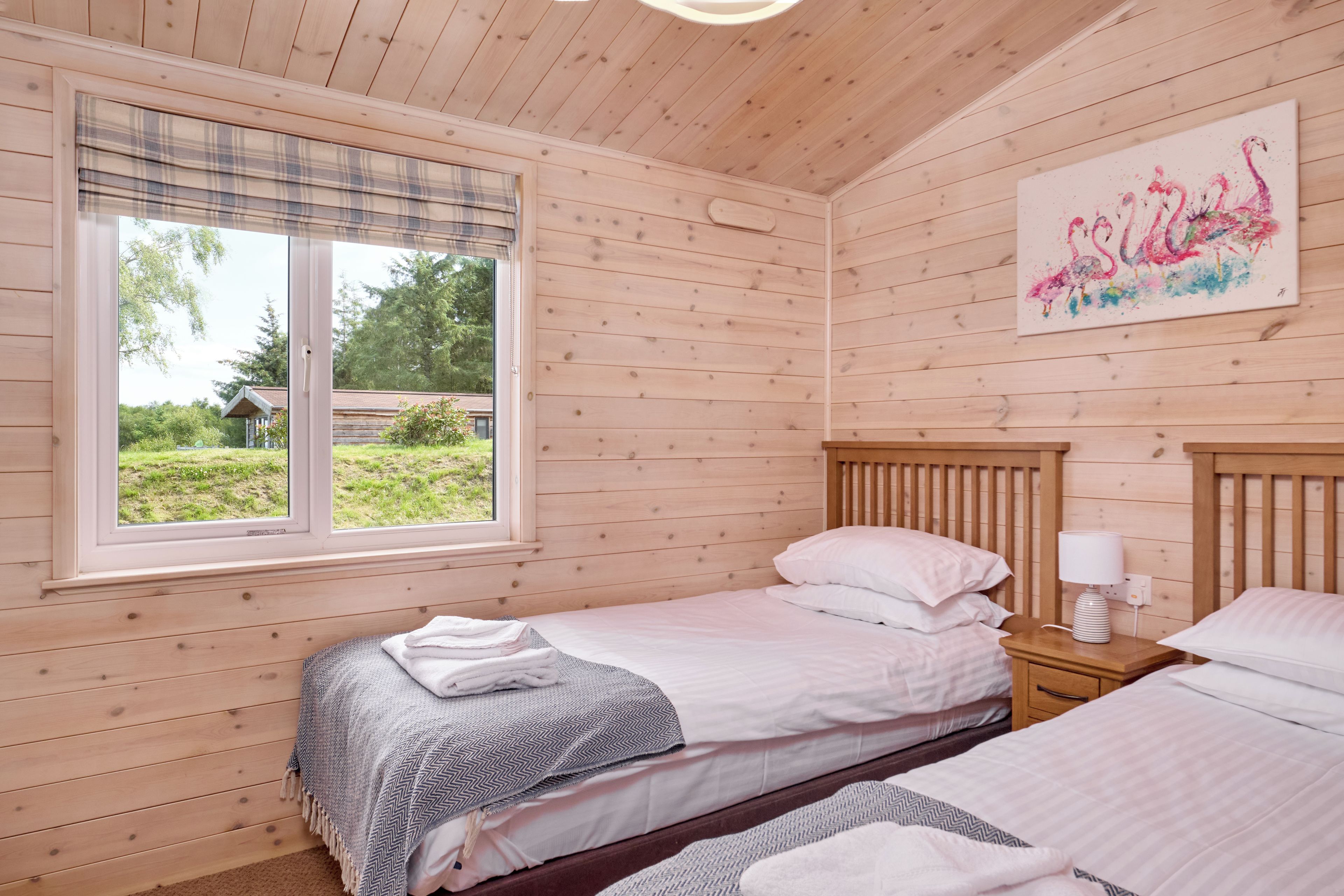 A wooden cabin room with two beds, a window with a view of greenery, and a painting on the wall.