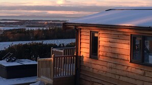 Une cabane en bois dotée d’une terrasse, entourée d’un paysage enneigé et offrant une vue sur des champs lointains et un ciel nuageux.