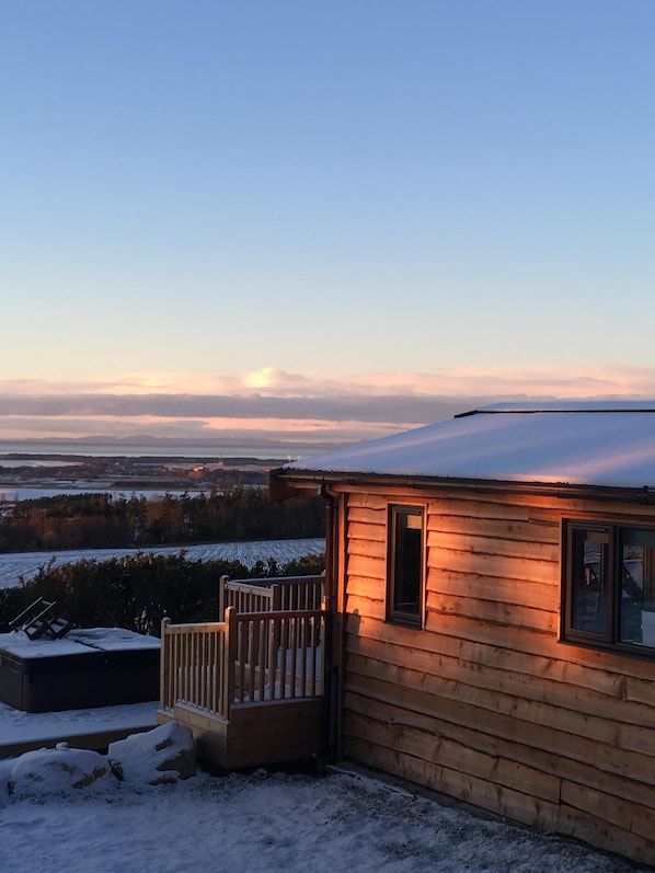 Una cabaña de madera con terraza, rodeada por un paisaje cubierto de nieve y vistas a campos lejanos y un cielo nublado.
